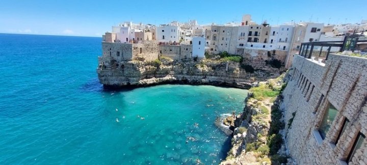 Domy nad onou „úžasnou“ zátokou v Polignano a mare. Azurově modrá a čistá voda a koupající se lidé všech generací. 