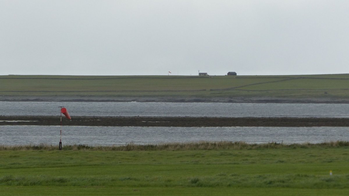 V popředí větrný rukáv letiště Westray. V pozadí terminál a větrný rukáv letiště Papa Westray